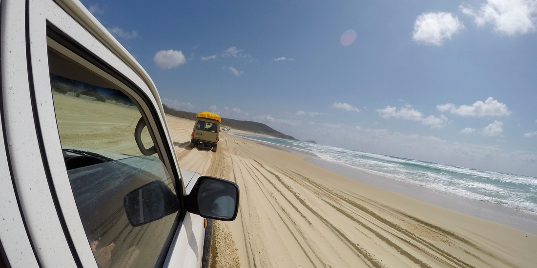 Vue depuis un 4x4 roulant sur une plage de sable en Australie, avec un autre véhicule en tête et l'océan en arrière-plan sous un ciel bleu clair